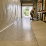 Gray flake floor coating installed in one zone of a Florida warehouse while plastic sheeting keeps the rest of the floor open
