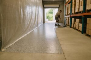 Gray flake floor coating installed in one zone of a Florida warehouse while plastic sheeting keeps the rest of the floor open